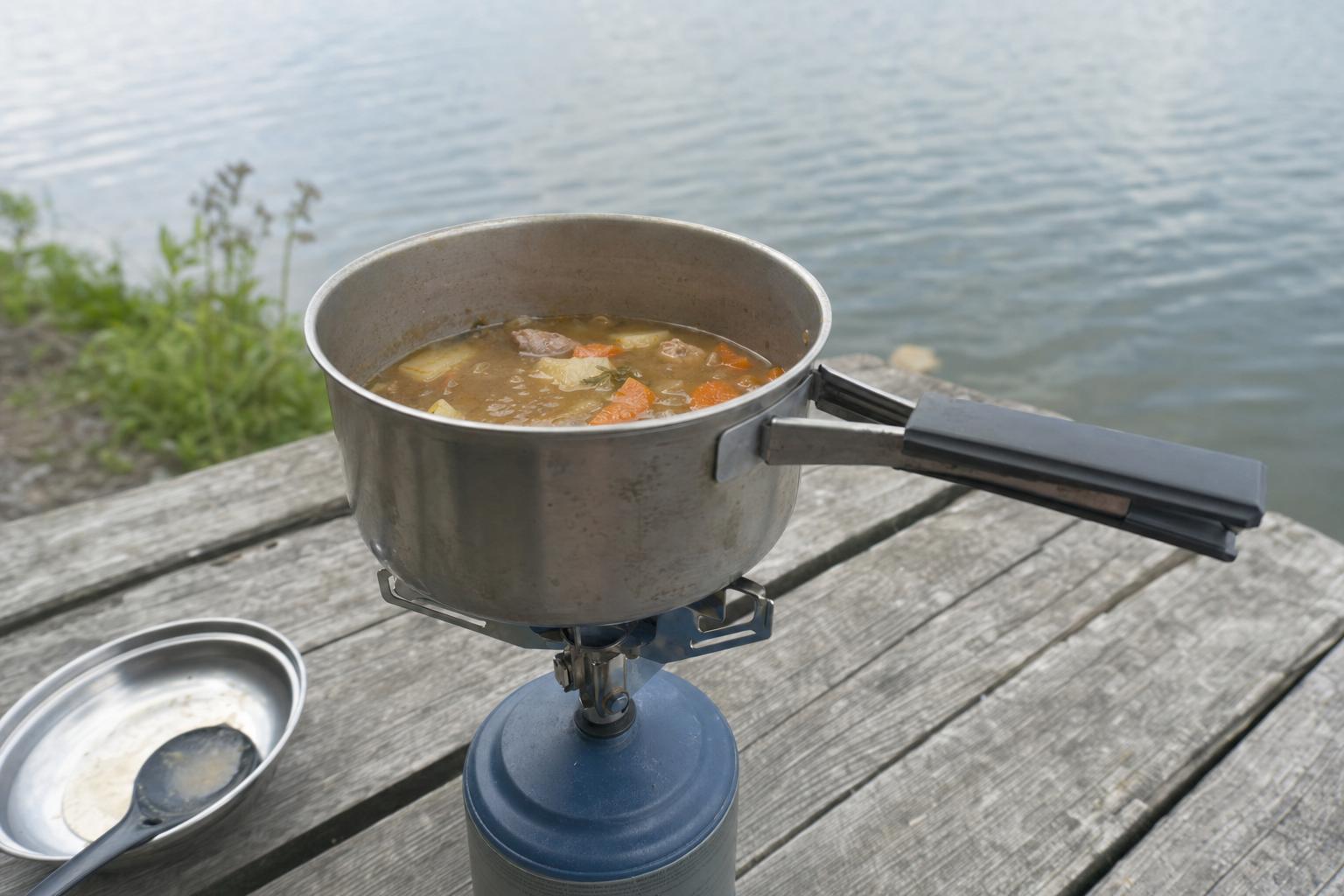A simple one-pot meal cooking on a small camp stove on a wooden table by a lake, no people