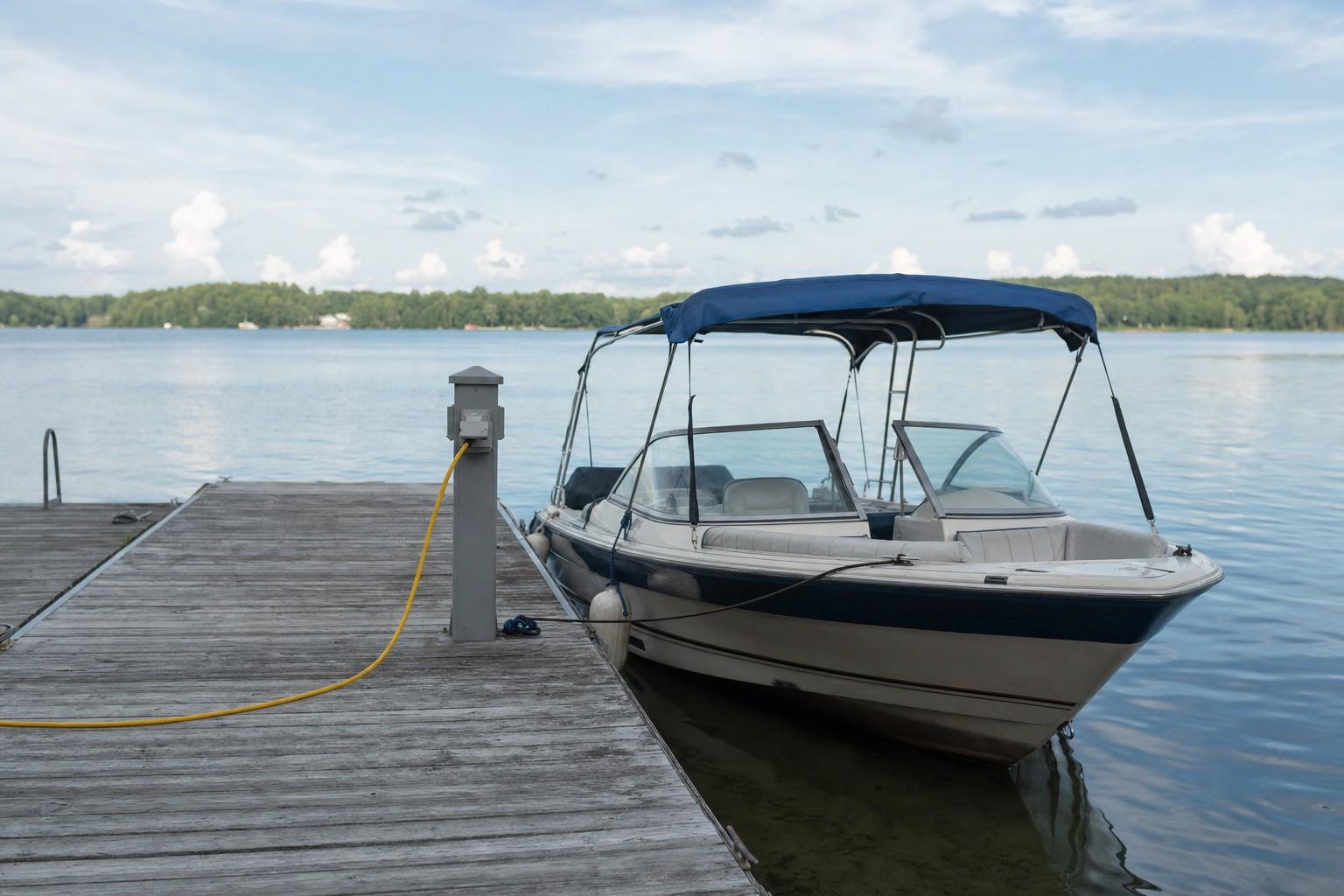 A small boat moored at a wooden lake dock with a shore-power pedestal on the deck