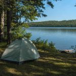 A small dome tent pitched on flat grassy ground under trees near a calm lake on a summer morning
