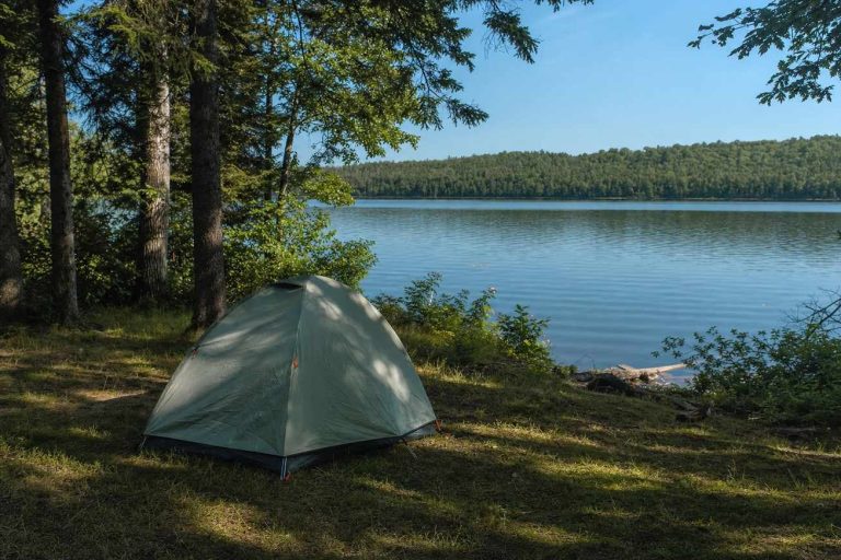 A small dome tent pitched on flat grassy ground under trees near a calm lake on a summer morning