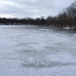 A wide view of a frozen lake with patchy snow showing uneven ice texture under an overcast winter sky