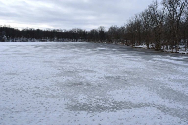A wide view of a frozen lake with patchy snow showing uneven ice texture under an overcast winter sky