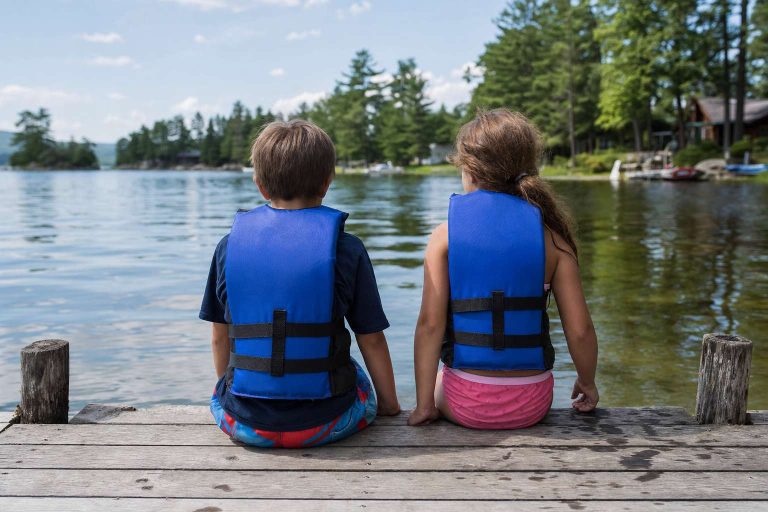 Two children in life jackets sitting on the edge of a dock with their feet over the calm water, seen from behind