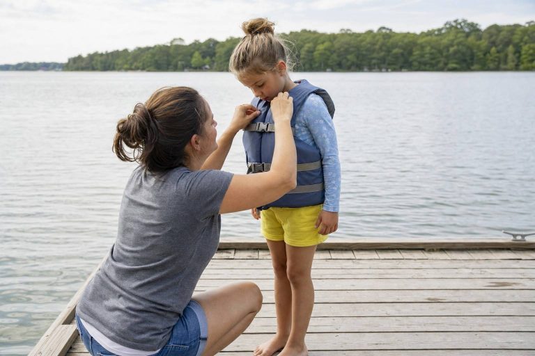An adult checking the strap of a child's life jacket on a dock by a calm lake