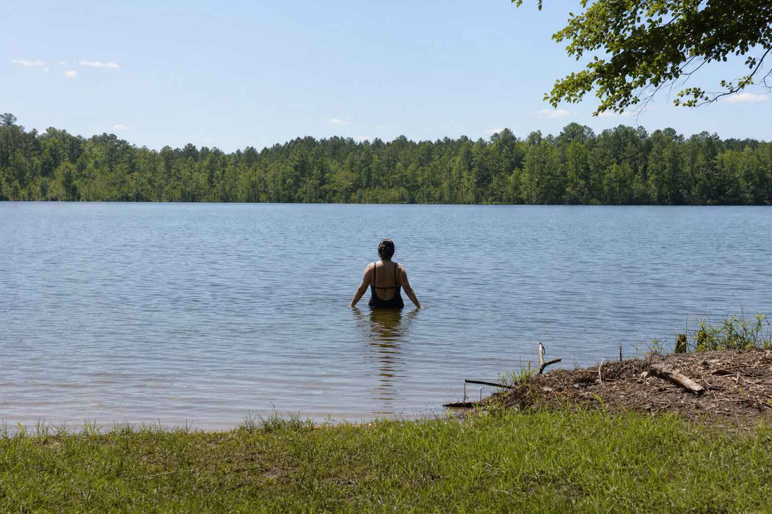 A person wading into a calm lake from a grassy shore, seen from behind, waist deep