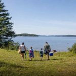 A small family arriving at a calm lake on a summer morning, carrying towels and a cooler toward a grassy shoreline
